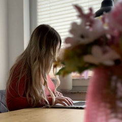 Eine Frau mit langen blonden Haaren arbeitet konzentriert an einem Laptop in einem gemütlichen, hellen Raum. Im Vordergrund sind unscharf Blumen zu sehen, die eine warme und kreative Atmosphäre schaffen.