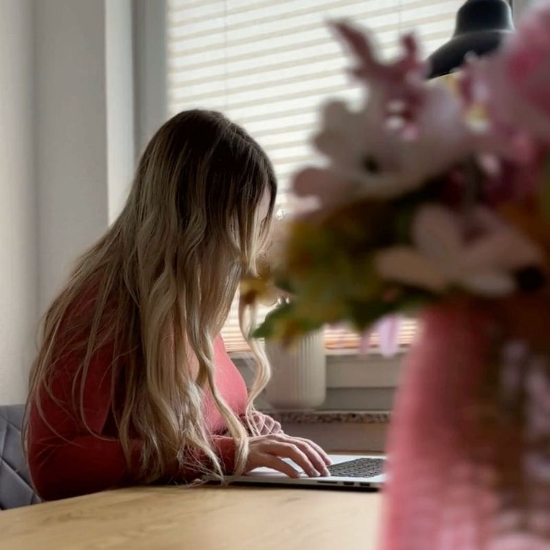 Eine Frau mit langen blonden Haaren arbeitet konzentriert an einem Laptop in einem gemütlichen, hellen Raum. Im Vordergrund sind unscharf Blumen zu sehen, die eine warme und kreative Atmosphäre schaffen.