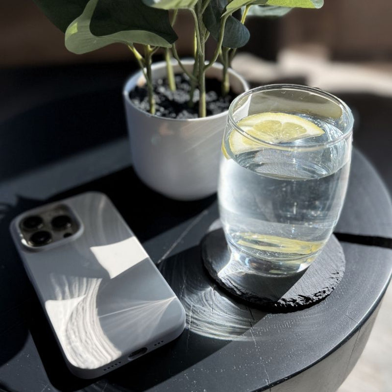 Ein minimalistischer Tisch mit einer Glaswasserkaraffe mit Zitronenscheibe, einem modernen Smartphone in einer weißen Hülle und einer kleinen Topfpflanze als Dekoration. Die Szene ist in natürlichem Licht gehalten und symbolisiert einen eleganten und organisierten Arbeitsplatz.